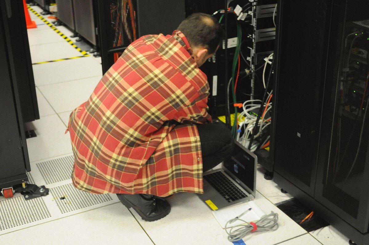 A technician kneeling at the back of an open server rack with a laptop on the raised floor, tracing cables by hand