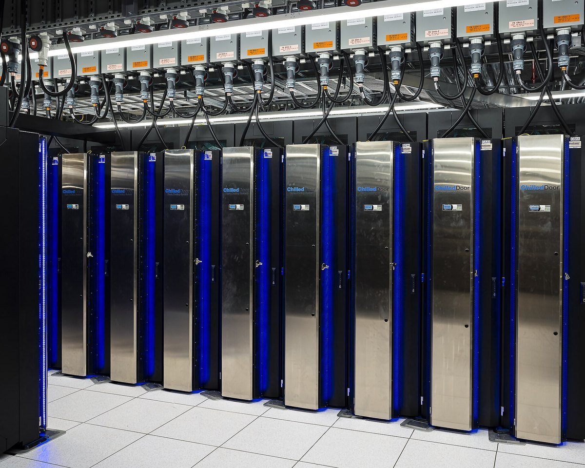 Overhead power whips with labelled circuit breakers feeding rack-door liquid cooling units in a supercomputer machine room