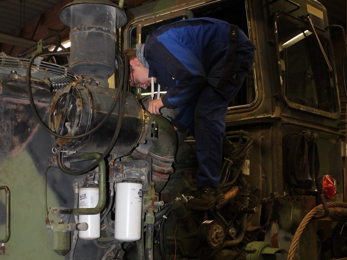 A maintenance apprentice in blue coveralls leaning over the engine bay of a military truck in a workshop