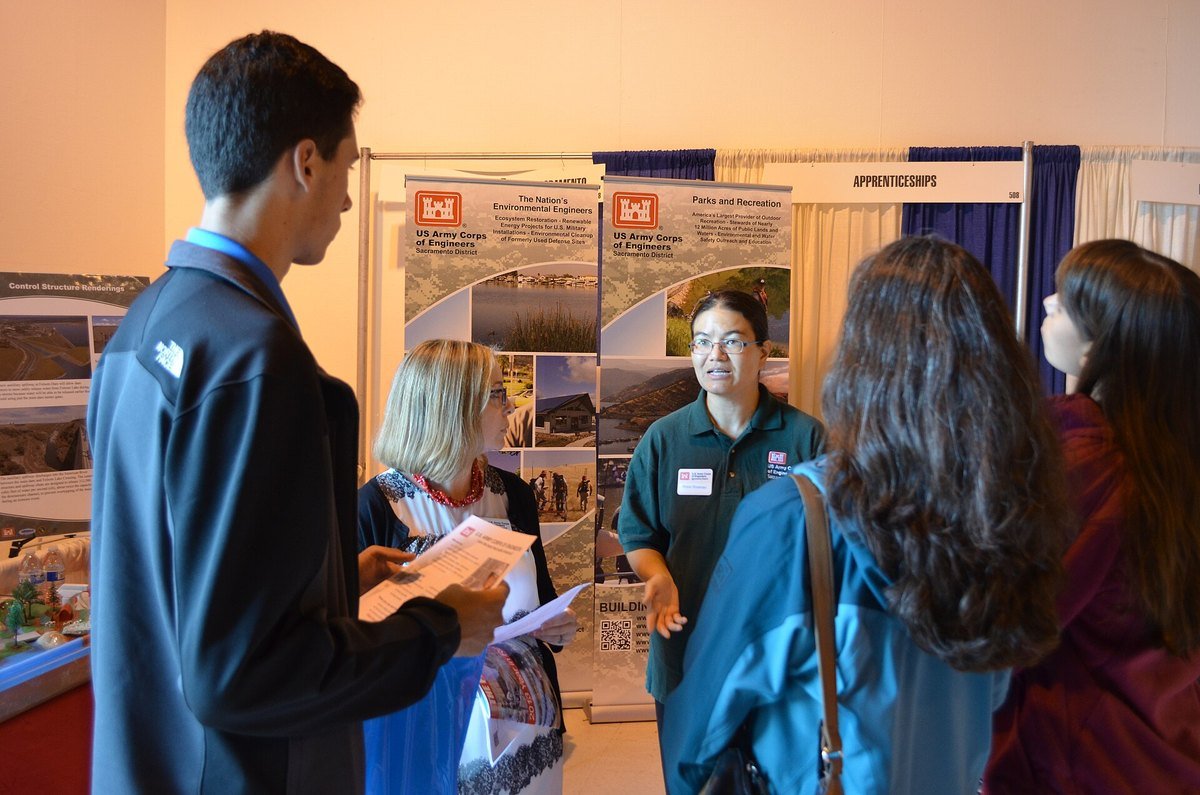 Job seekers talking with recruiters at booths during a public engineering career fair, with an Apprenticeships sign visible above the aisle