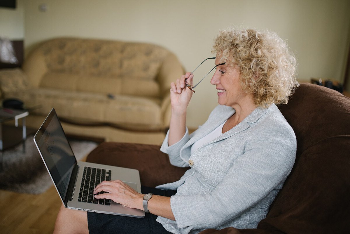 An older woman sitting at home with a laptop, glasses in hand