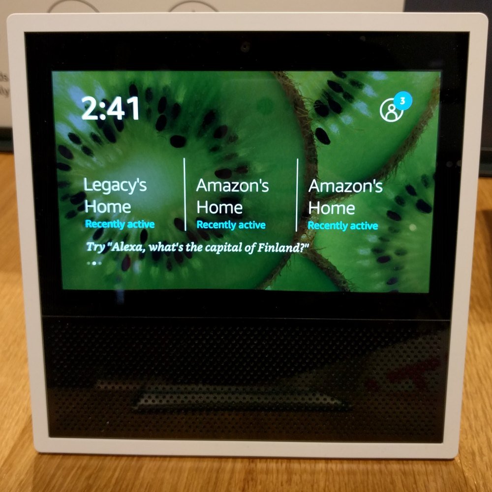 An Amazon Echo Show displaying a rotating home screen card that reads Try Alexa, what's the capital of Finland
