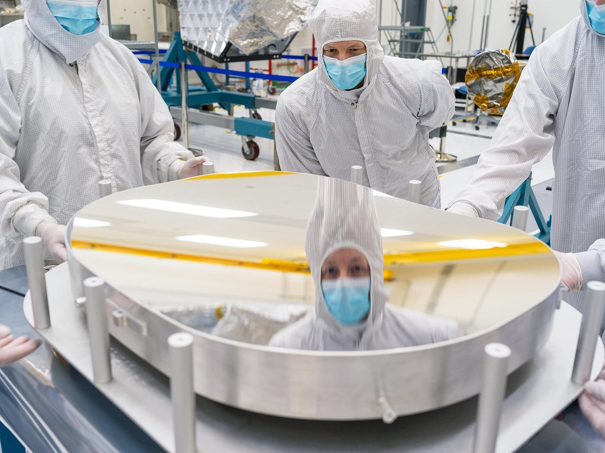 Technicians in white cleanroom suits and masks inspecting a large circular telescope mirror at a federal laboratory