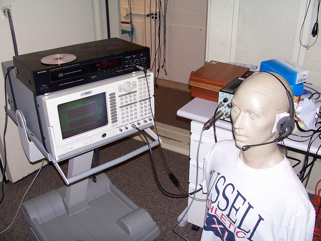 A call centre headset fitted to a mannequin test head wired to laboratory measuring equipment.