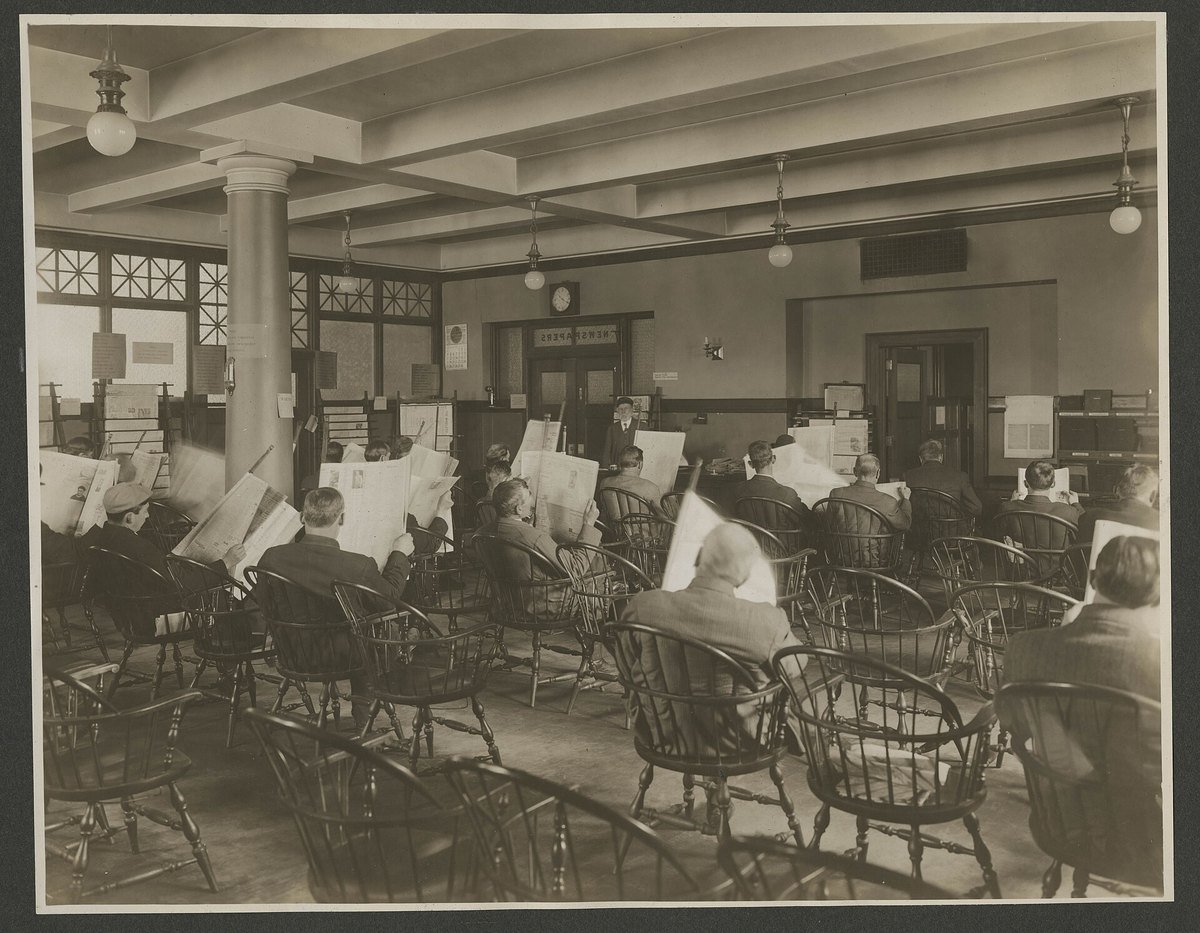 Men reading newspapers in the Seattle Central Library newspaper room around 1910, under a sign reading NEWSPAPERS