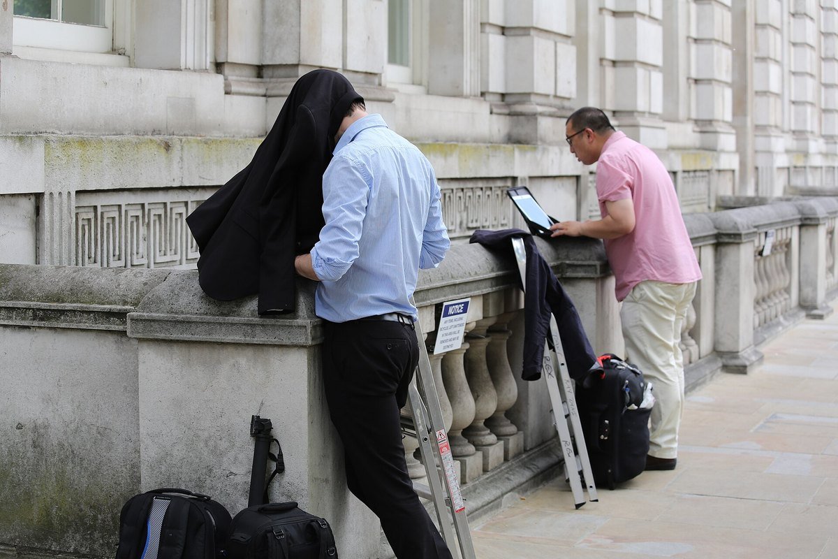 Two press photographers filing pictures from the street outside a government building, one shielding a screen with a jacket