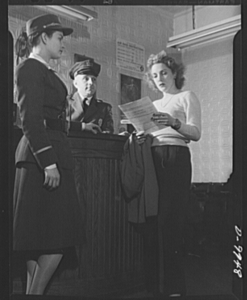 A woman reads a job application form at a plant gate while two guards look on, 1943