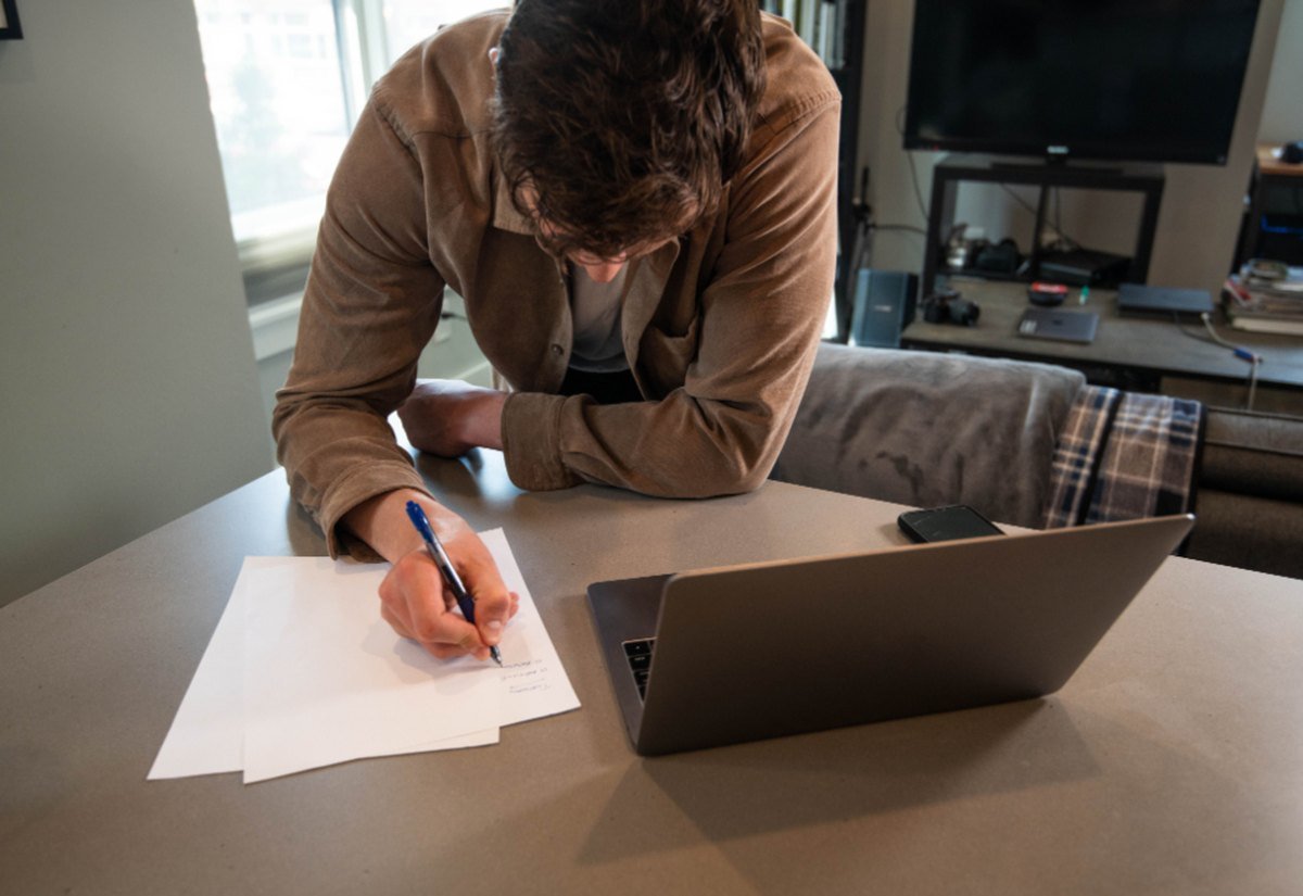 A person standing at a kitchen table with a laptop open and paperwork spread out, writing