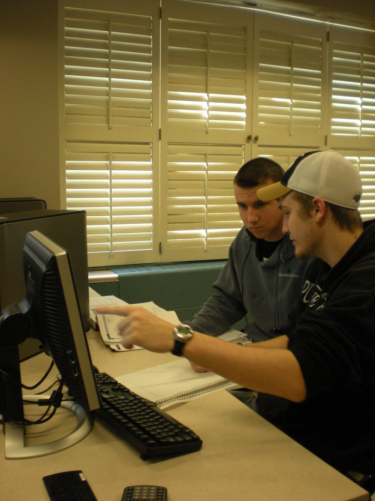 Two students working together at a school computer with notebooks and a calculator