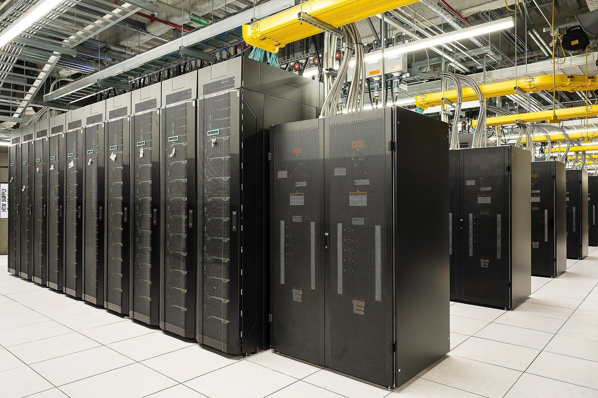 Rows of black server cabinets in a supercomputer machine room with overhead yellow cable trays and coolant piping