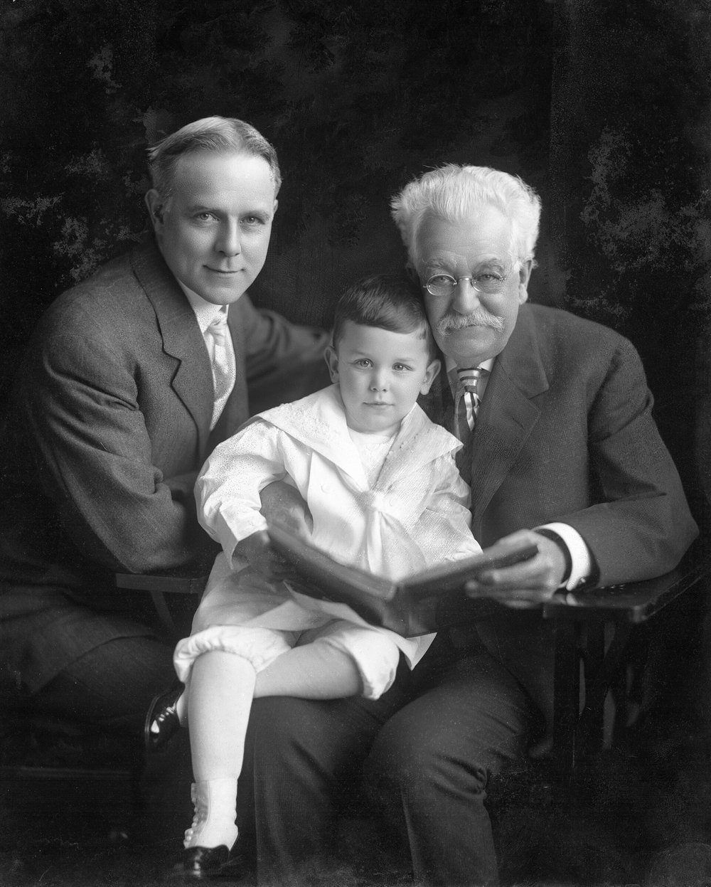 A grandfather, his son and a small grandchild sharing an open book in a studio portrait, about 1920