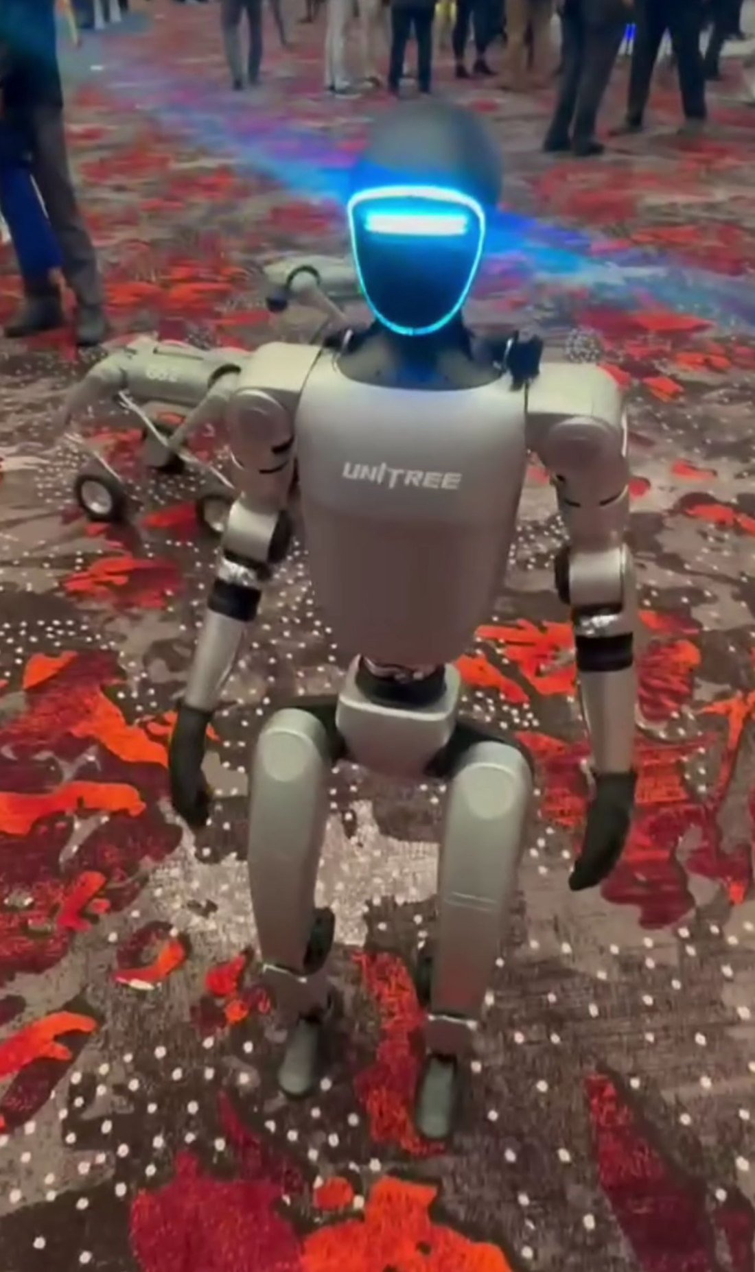 A Unitree G1 humanoid robot standing on display at an expo, showing its jointed arms, torso and head sensor array.