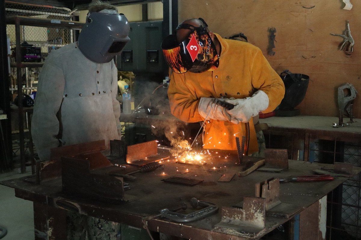 Two welders in protective gear at a workbench with sparks flying during a basic welding class