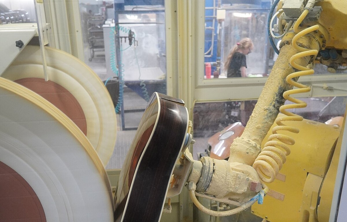 A dust-covered yellow industrial robot arm holding a guitar body against a spinning buffing wheel inside a safety enclosure, with a worker visible through the glass behind it
