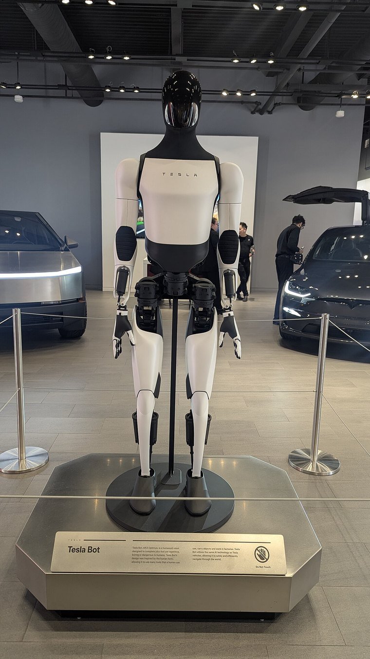 A white and black Tesla Optimus humanoid robot standing on a display plinth inside a Tesla showroom, with cars in the background