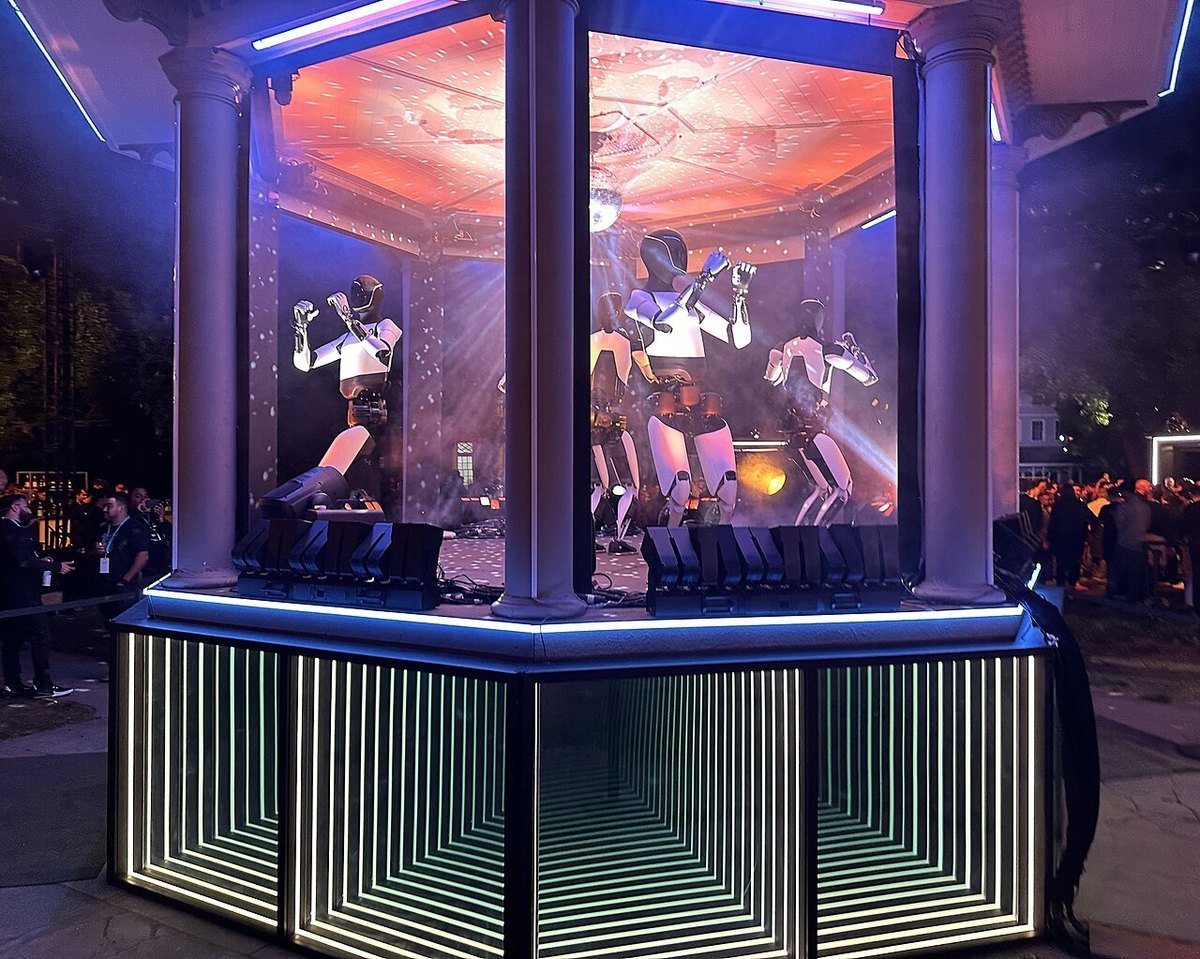 Several Tesla Optimus robots dancing inside a lit glass gazebo at night while a crowd watches