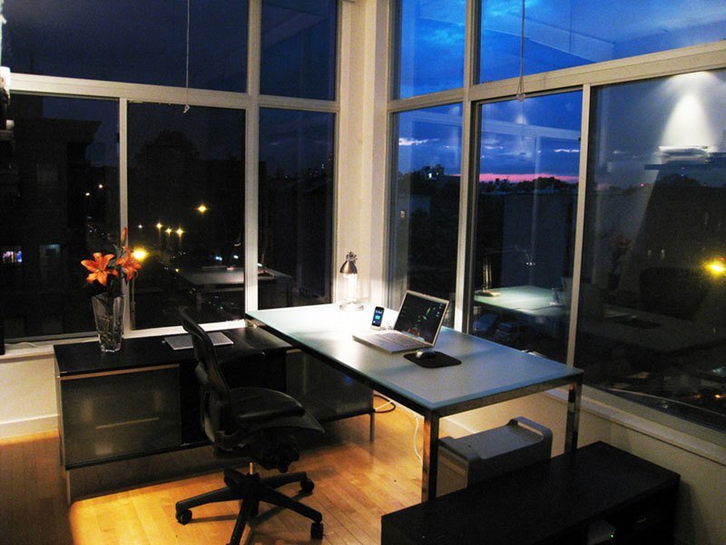 Home office desk with a laptop beside tall windows overlooking city lights at dusk