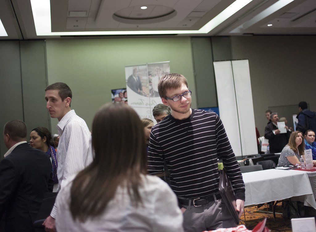 Job seekers walking between employer tables at an indoor career fair, several carrying folders