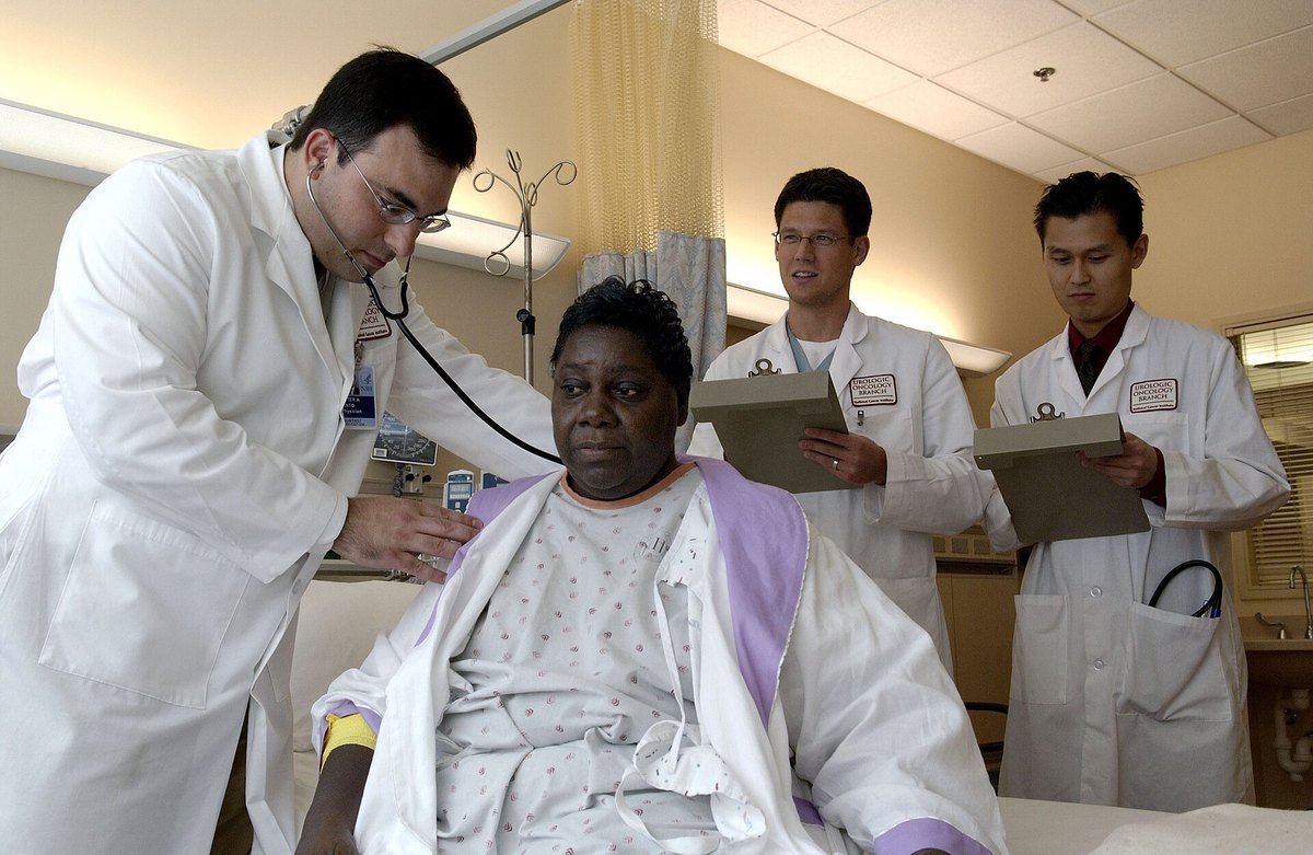 A physician examines a patient while two other clinicians stand nearby holding chart folders and clipboards
