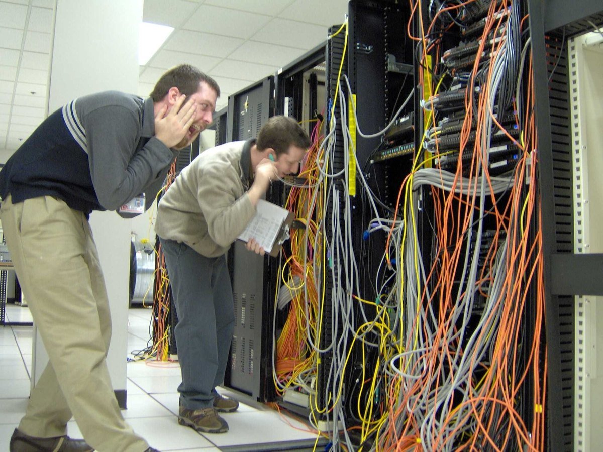 Two data center operators leaning into a rack, tracing a dense tangle of orange and grey network cables.