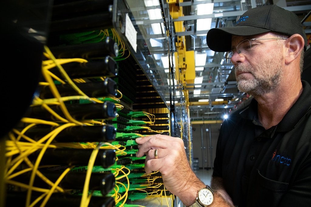 Technician in a cap and glasses working on a rack of yellow and green fibre-optic patch cables inside a network facility