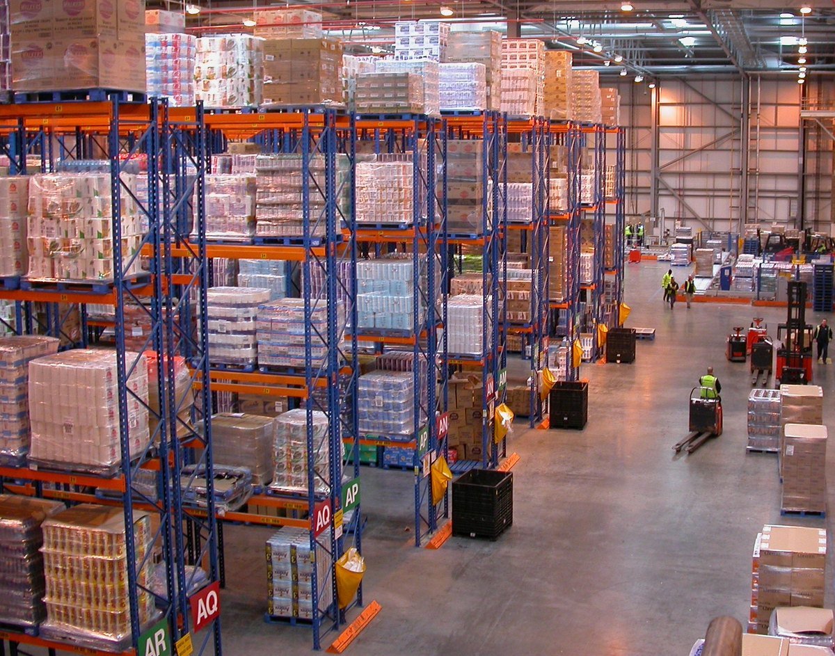 Interior of a large grocery distribution centre with pallet racking, aisle markers and workers on pallet trucks.
