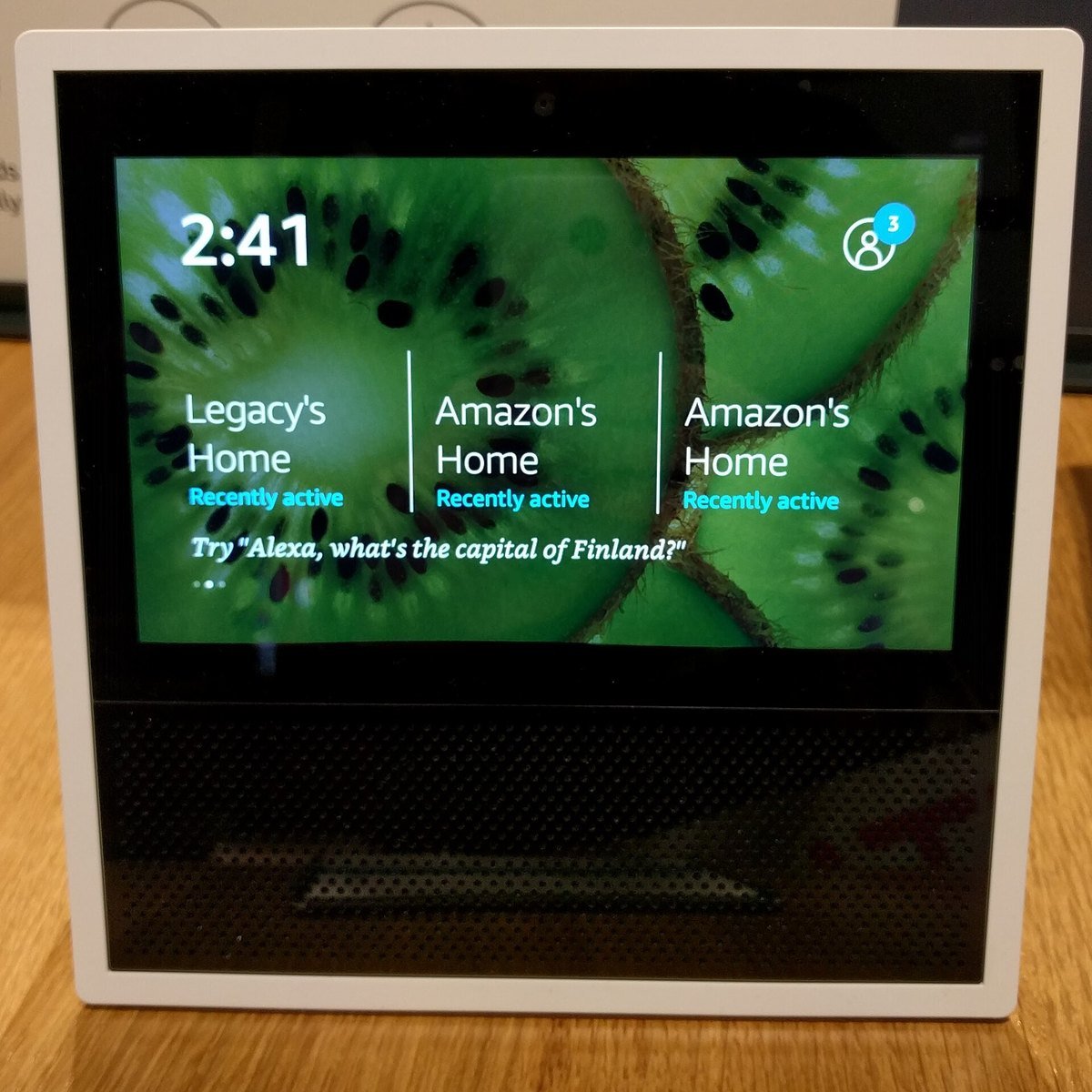 An Amazon Echo Show on a wooden table displaying suggestion cards and a prompt reading Try Alexa, what is the capital of Finland