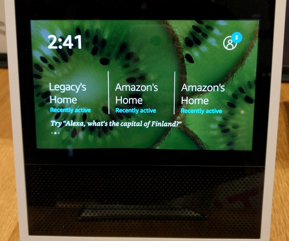 A first-generation Amazon Echo Show smart display in white showing a clock and household names on screen
