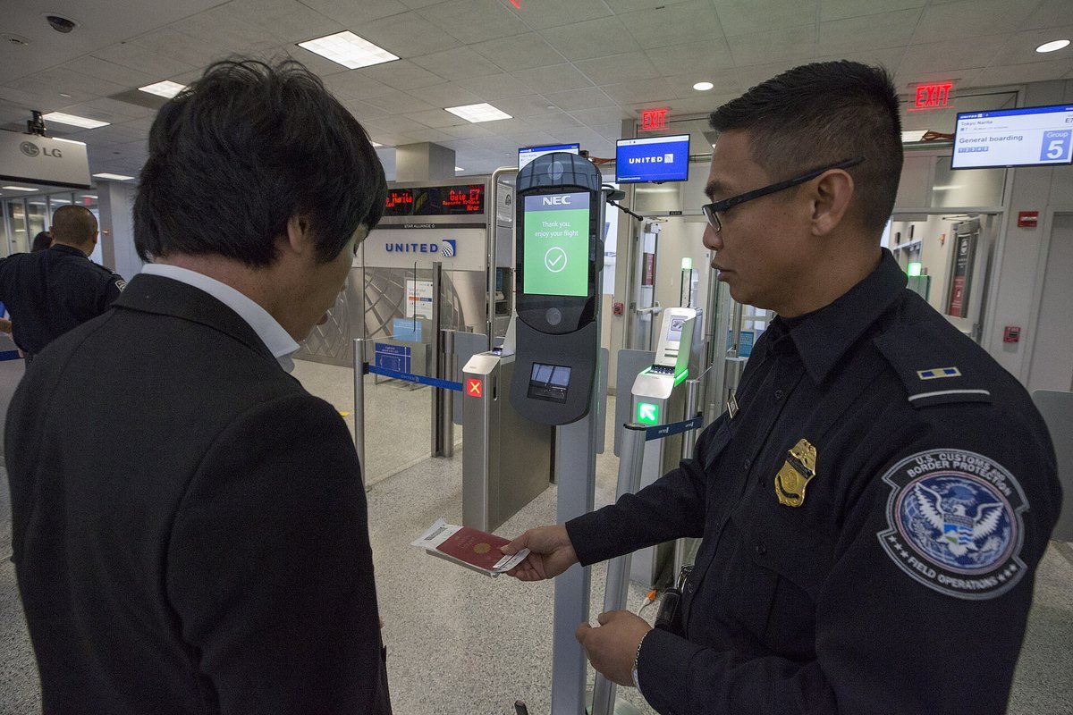 A traveller at an airport gate facing a camera kiosk while an officer checks a passport.
