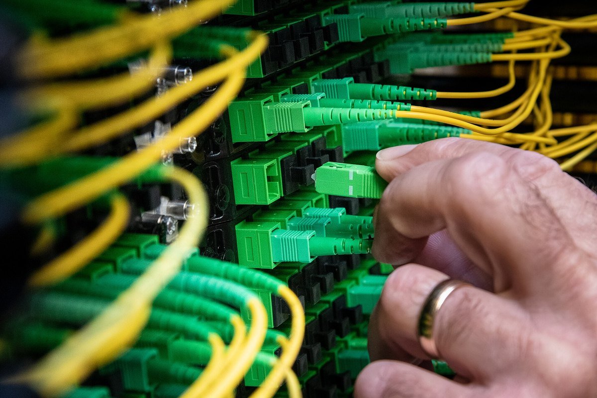 Close-up of a technician's hand seating a green fibre-optic connector into a dense patch panel of yellow and green cables.
