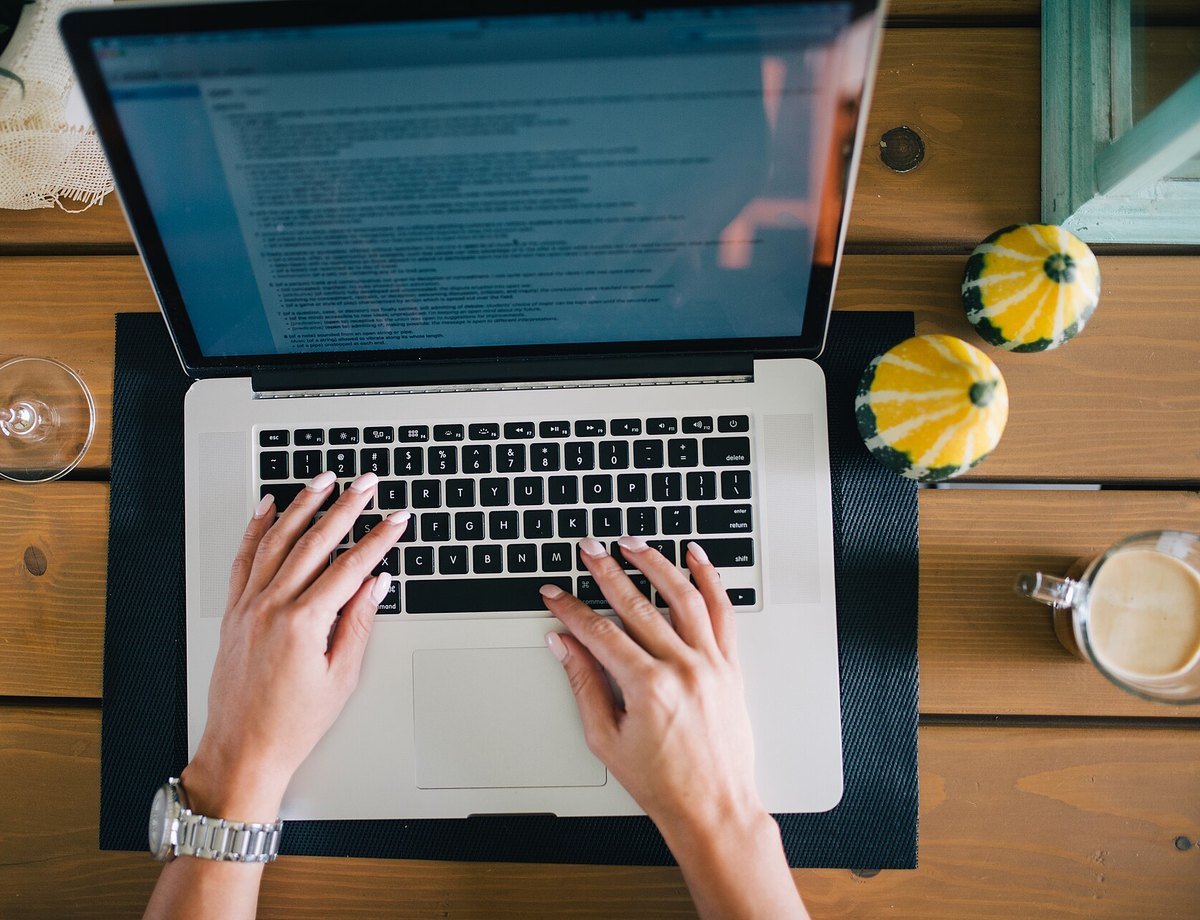 Overhead view of hands typing on a laptop at a wooden kitchen table with a coffee beside it.