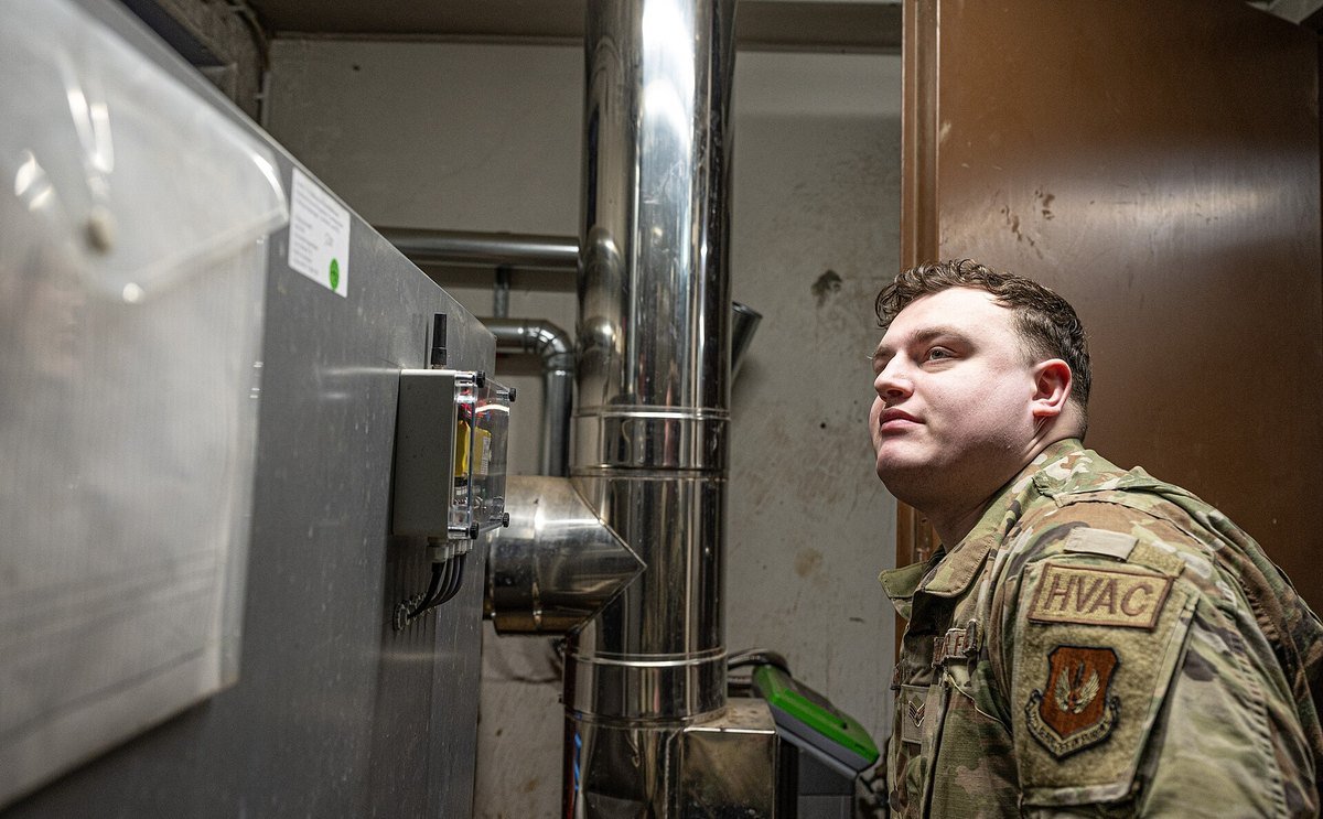 A technician in uniform with an HVAC patch on the sleeve inspecting a metal air-handling unit and ductwork in a mechanical room.