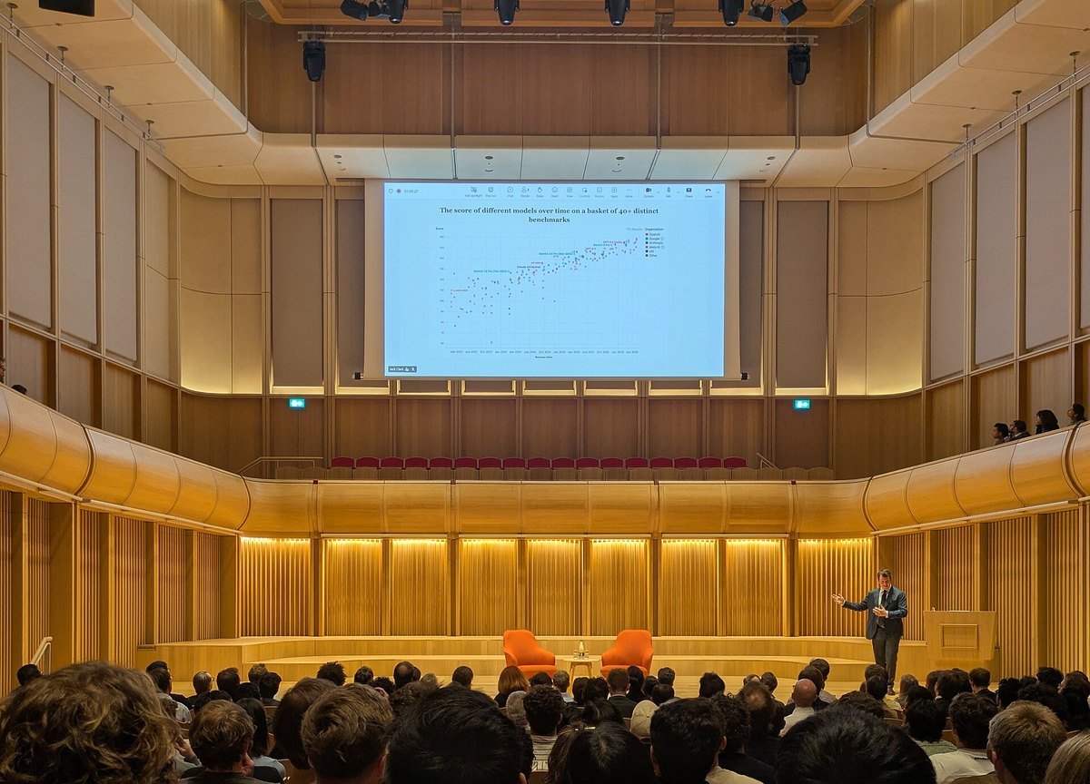 A speaker on stage in a wood-paneled concert hall presenting a chart of AI model benchmark scores to a large audience.
