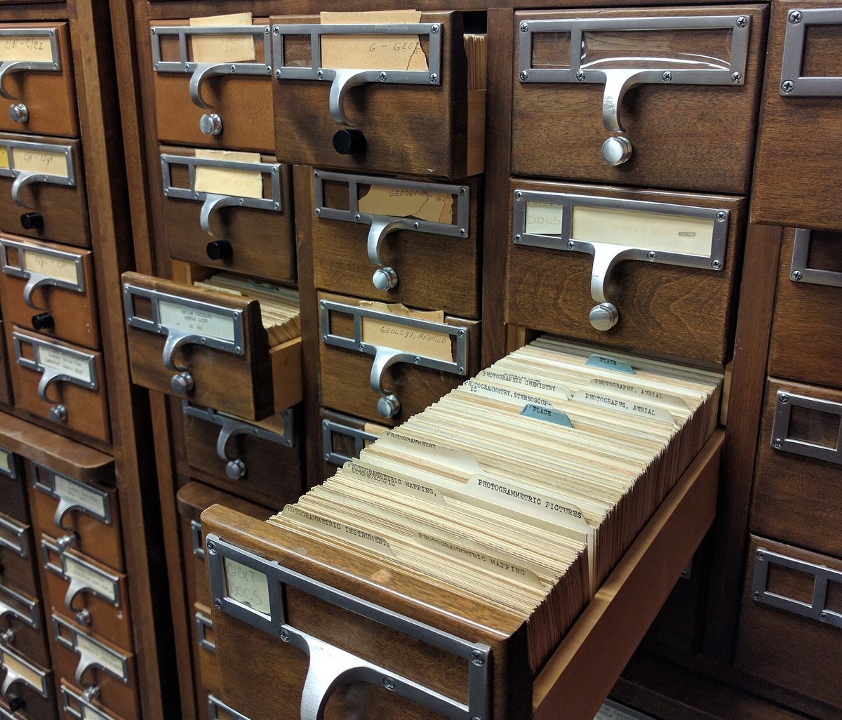 Wooden drawers of a library card catalog, one pulled open to show tightly packed index cards with subject headings.