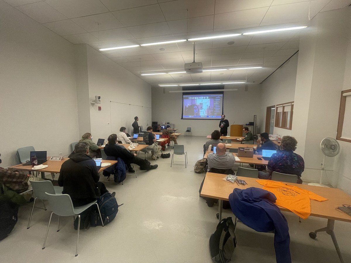A public library meeting room with a Zoom video call on a projector screen and people seated at tables with laptops