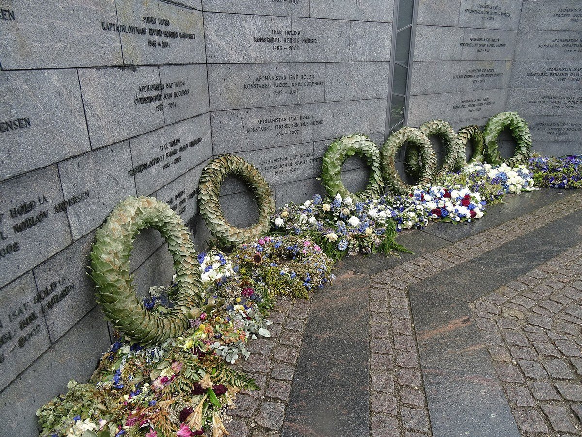 Funeral wreaths and bouquets lined along a stone memorial wall engraved with names.