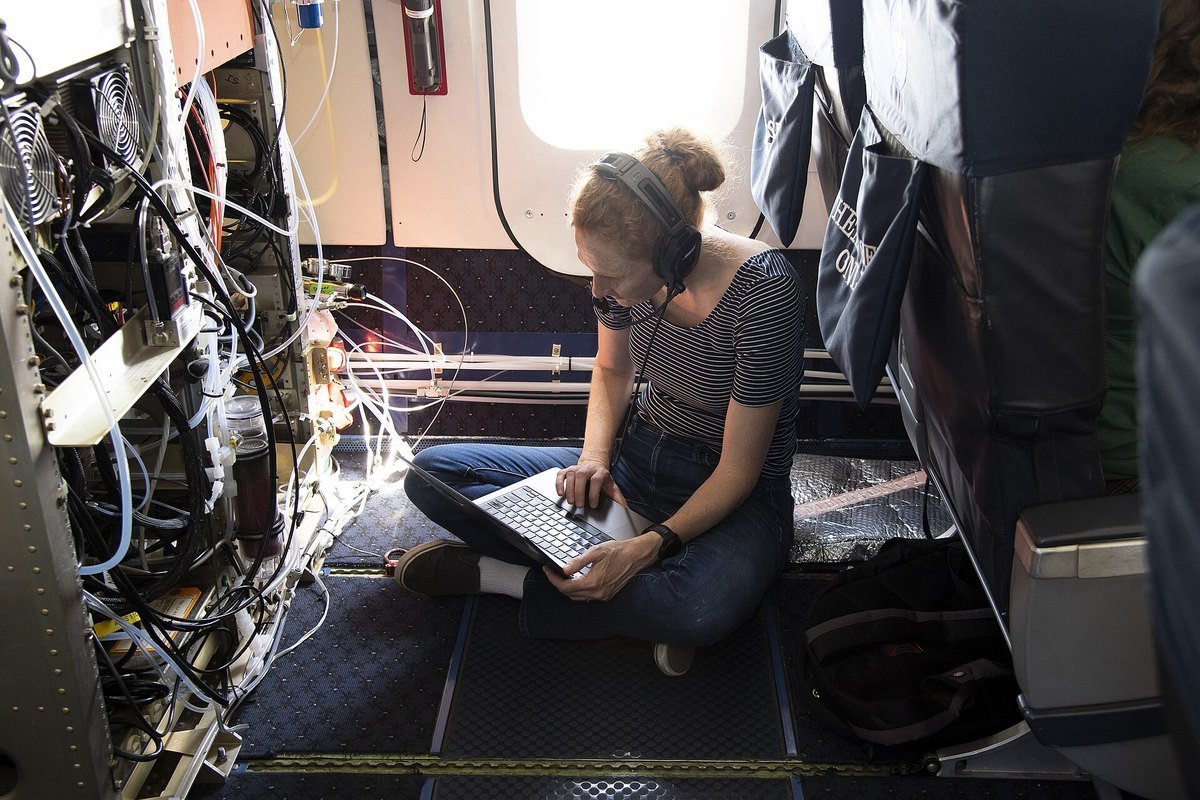 A student intern sits cross-legged on the floor of a research aircraft with a laptop, beside racks of scientific instruments
