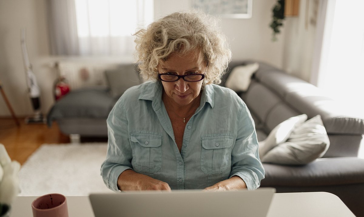 An older adult in a light blue shirt working on a laptop at a table in a living room.