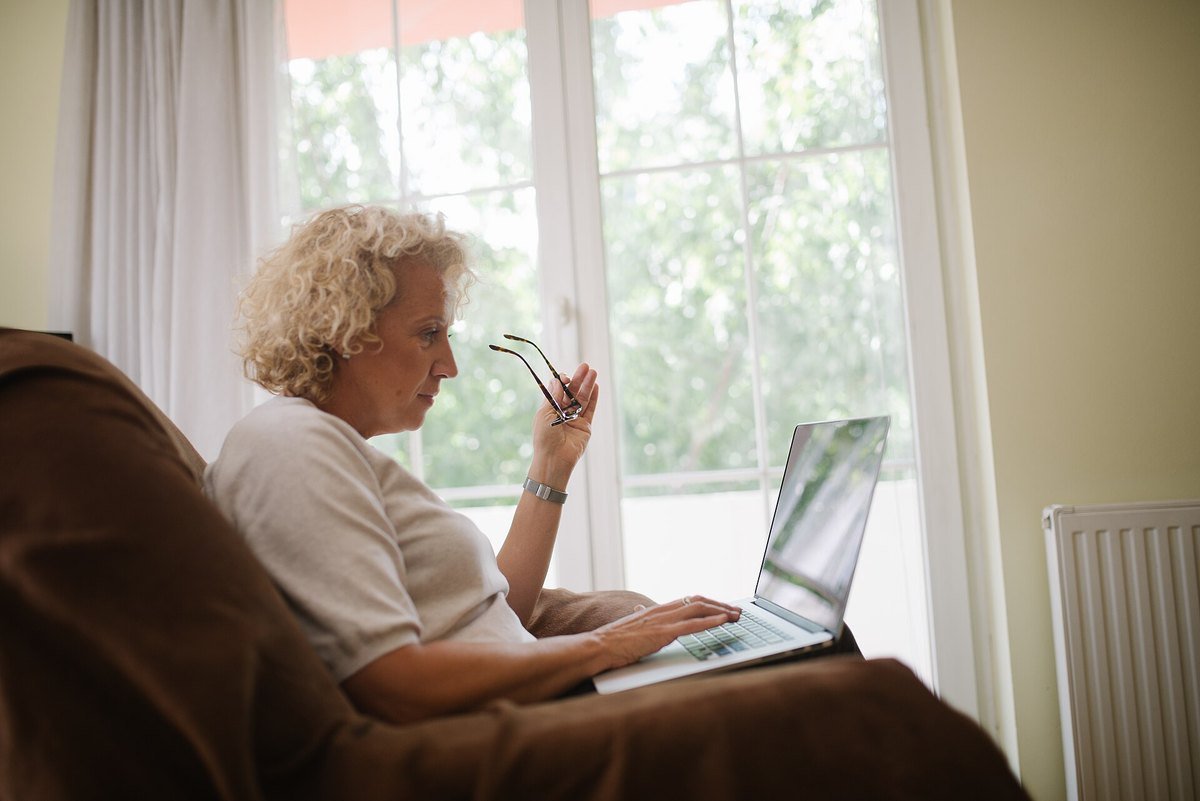 An older woman sitting in an armchair, holding her glasses while reading a laptop screen