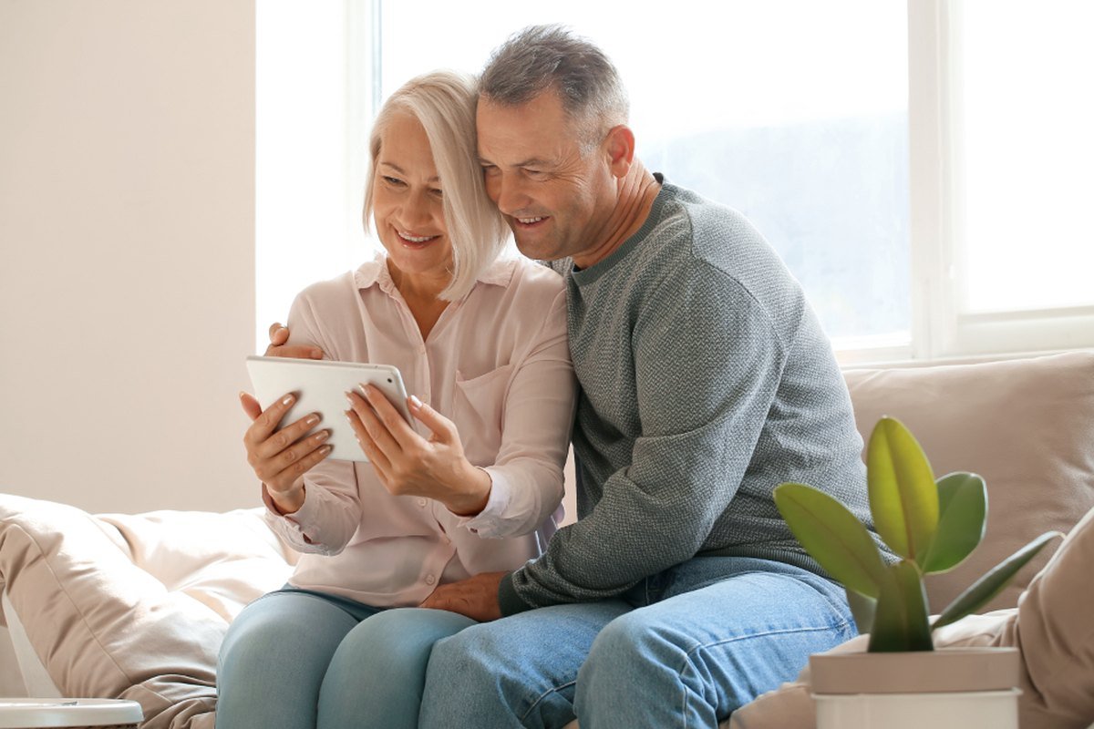 An older couple sitting on a couch by a window, looking at a tablet together.