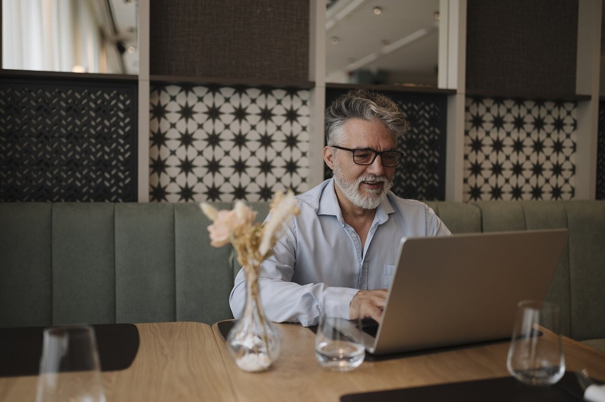 An older man with glasses smiling while using a laptop at a cafe table.