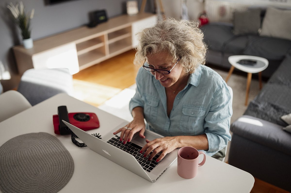 An older woman with glasses typing on a laptop at her kitchen table.