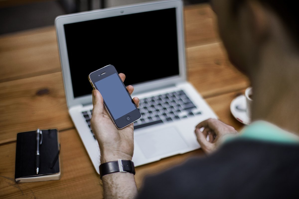 A person holding a smartphone while sitting at an open laptop