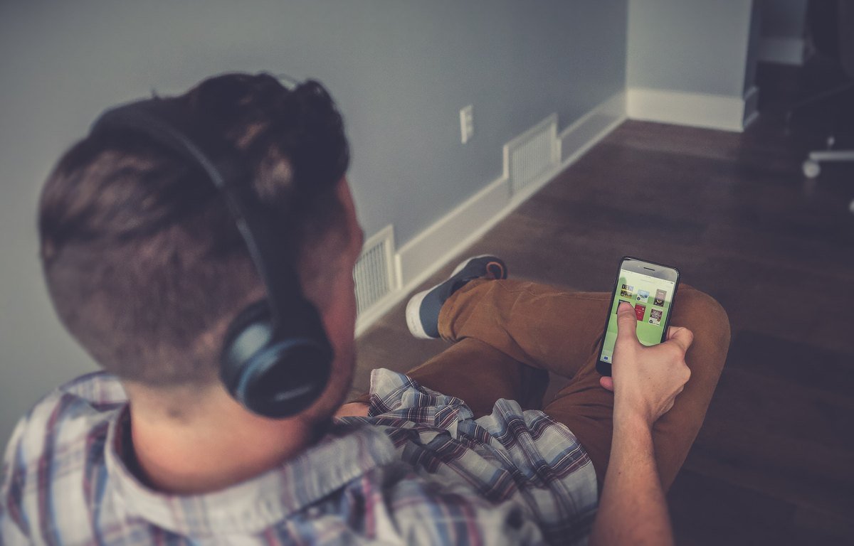 A man wearing headphones looks down at a smartphone in his hand, seen from over his shoulder.