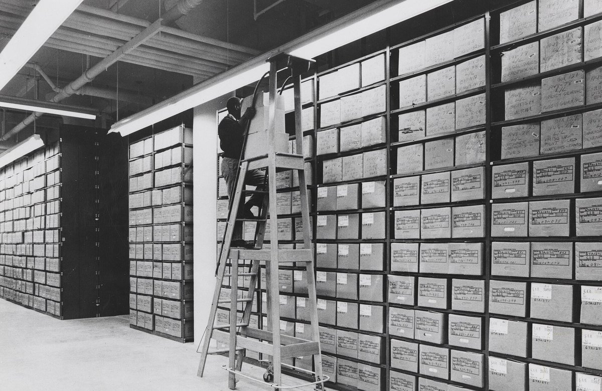 An archivist on a rolling ladder retrieves a records box from floor-to-ceiling shelves at the Washington National Records Center