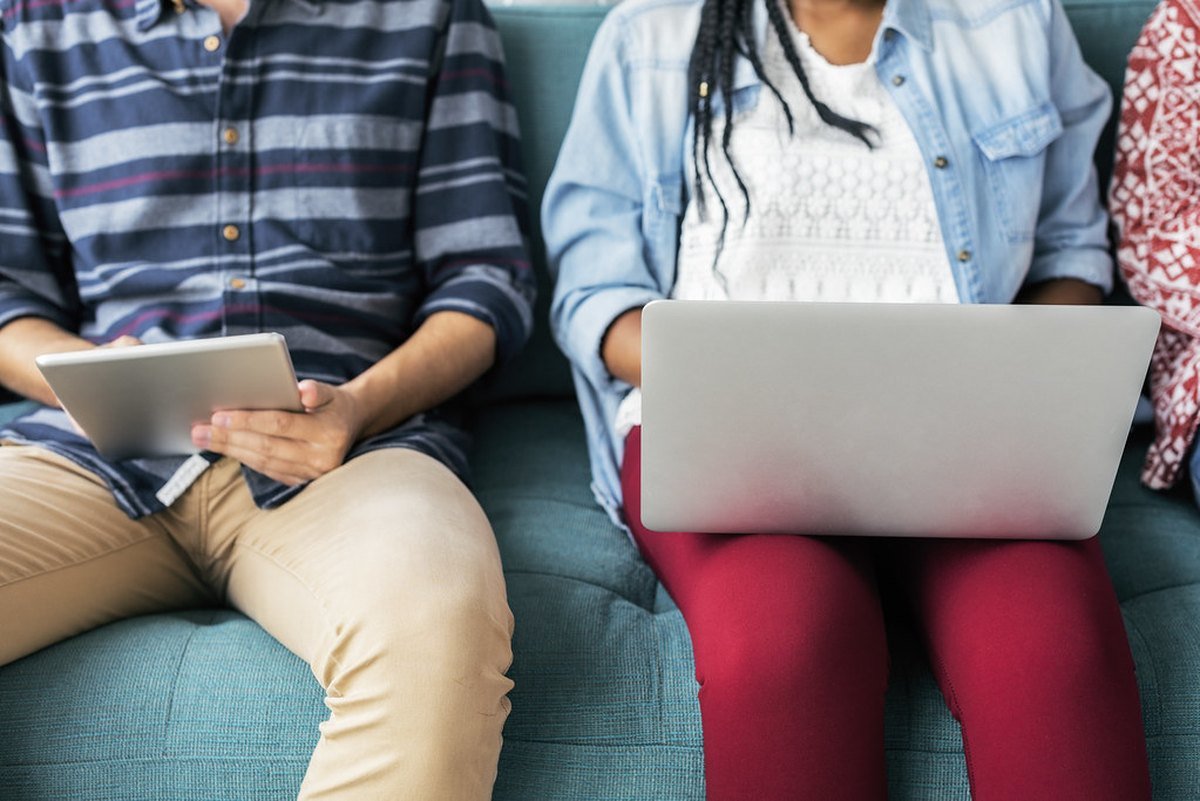 Two people sitting side by side on a couch, one holding a tablet and one a laptop.