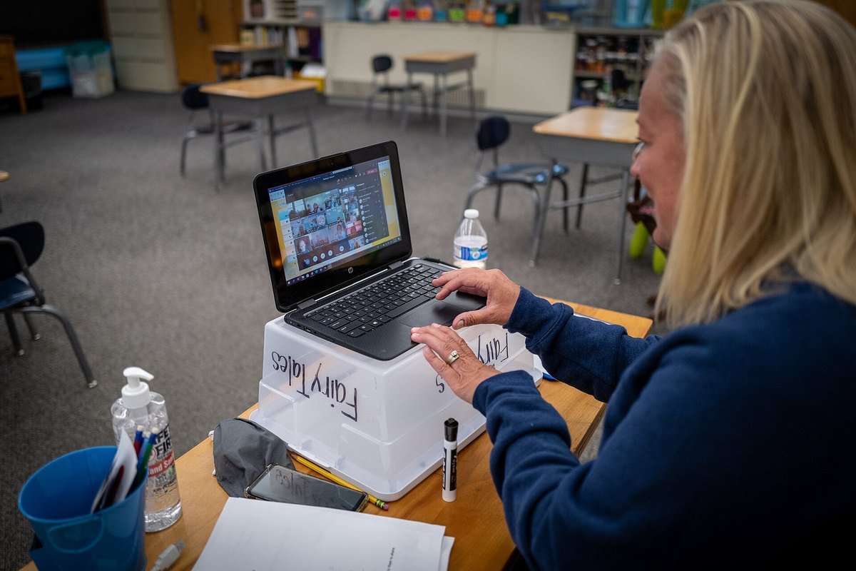 A teacher at a classroom desk delivering a lesson to students on a Zoom call on her laptop