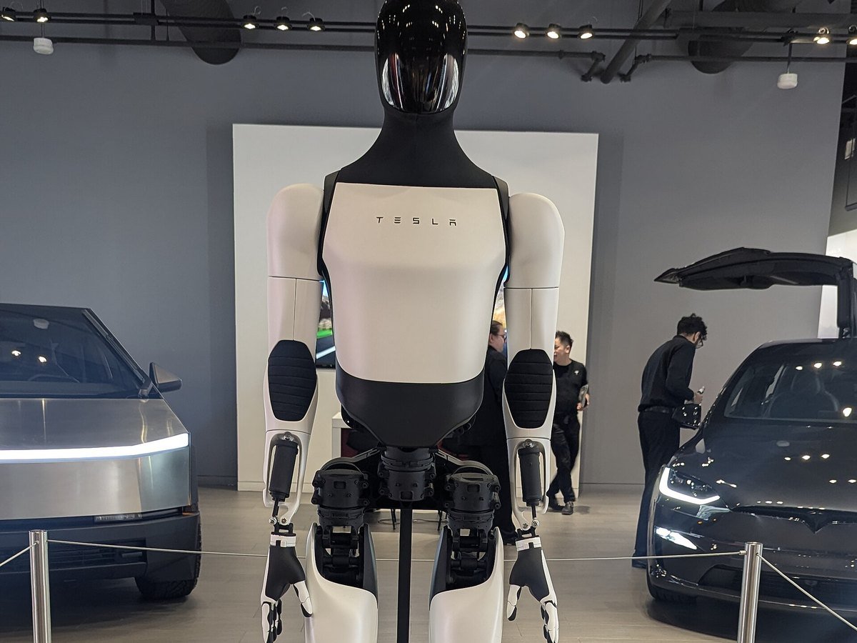 Tesla Optimus humanoid robot displayed between two cars in a showroom