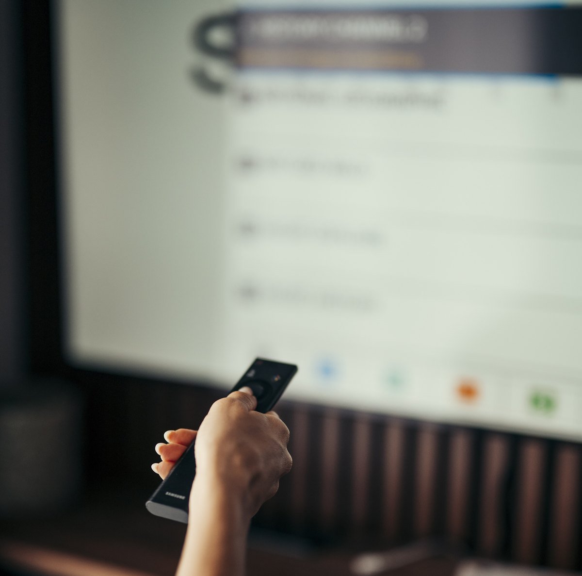 A hand points a TV remote at a large screen showing a blurred on-screen menu.
