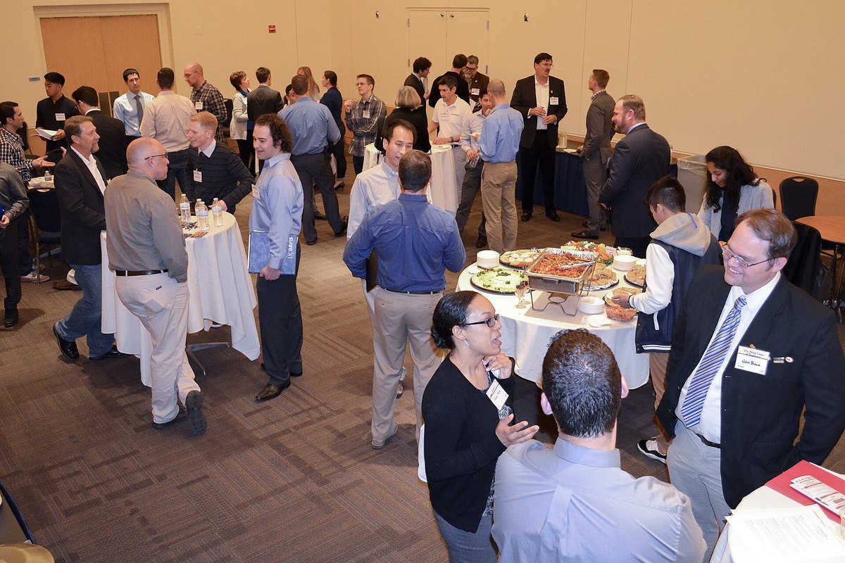 Students and employer representatives talking around tables at a crowded university job fair in a carpeted hall.